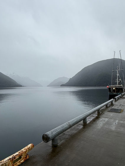 Rainy waterfront in Sitka, Alaska, overlooking a calm fjord with forested mountains and a commercial fishing vessel, representing the challenging maritime environments where Tiamat Maritime conducts survey and inspection operations.