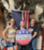 Three women volunteers holding a Packages From Home sign in front of an American flag.