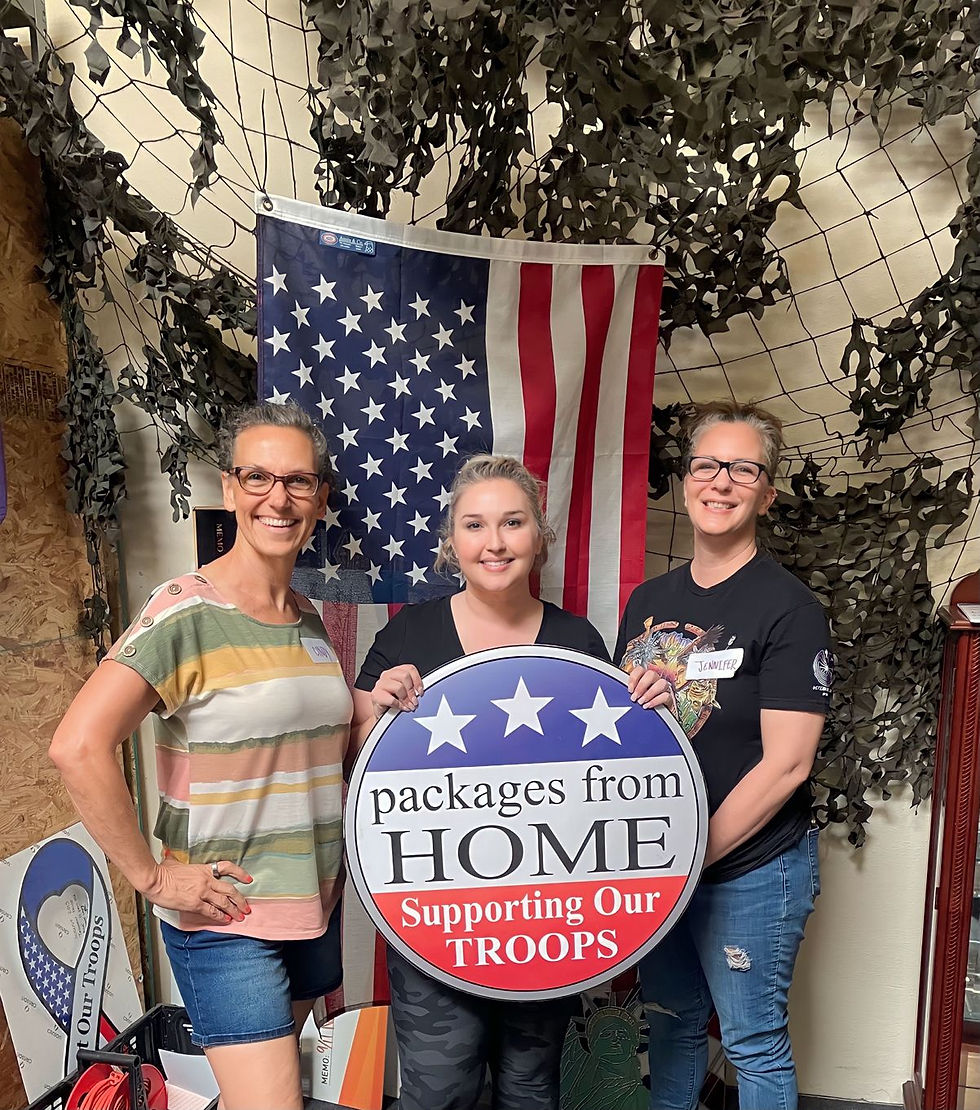 Three women volunteers holding a Packages From Home sign in front of an American flag.