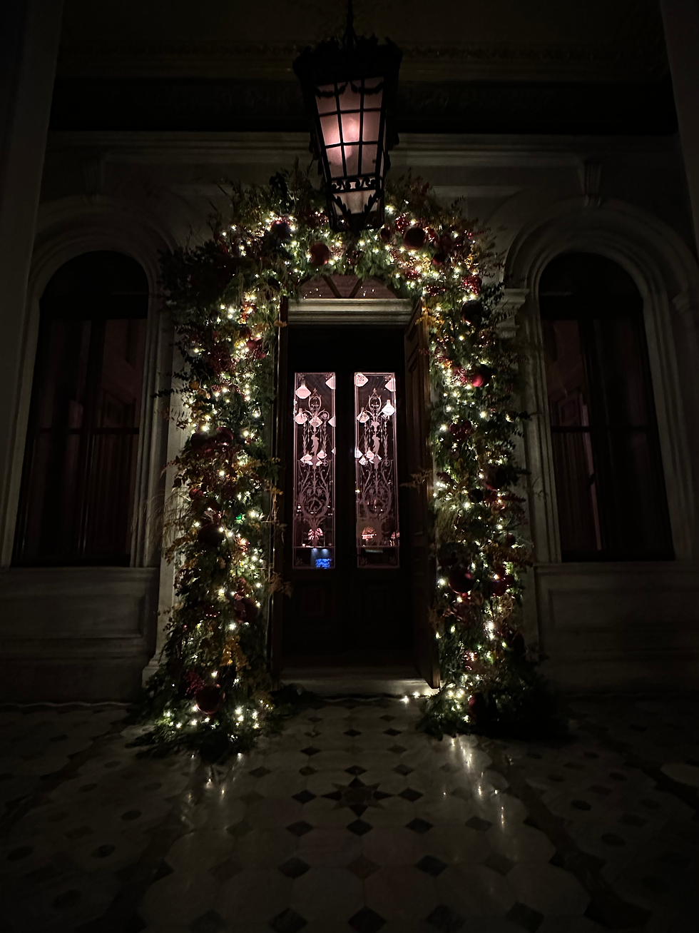 Dim doorway framed by glowing Christmas garland, red ornaments, and a hanging lantern in a dark hallway