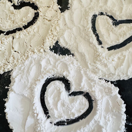 Flour spread on a dark surface forms three heart shapes. The setting is simple, with a playful and creative mood.