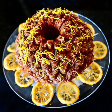 Limoncello Bundt cake topped with lemon zest, surrounded by lemon slices on a glass plate. Dark background, bright and vibrant presentation.