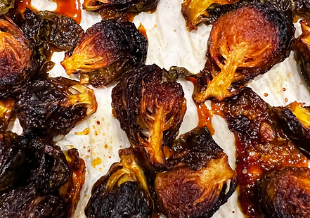 Crispy, roasted Brussels sprouts, halved and tossed in a glossy, sticky red gochujang glaze. The dish is garnished with toasted sesame seeds and thinly sliced green onions (or cilantro/peanuts), served in a bowl or on a platter.
