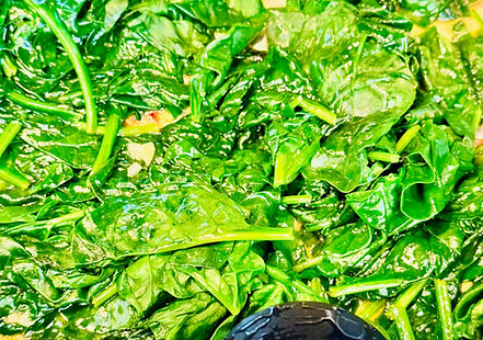 Sautéed spinach with a glossy finish, cooked in a pan, with a black slotted spoon partially visible. The dish appears fresh and vibrant.