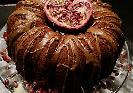 A rich, moist, gluten-free bundt cake with hints of honey and apple sits on a white serving platter. The cake is generously draped in a sweet cinnamon glaze that cascades down its ridges, pooling slightly at the base. The glaze is dusted with additional ground cinnamon, and the cake is garnished with a few thinly sliced, fresh red apples arranged at the base of the platter, adding a pop of vibrant color. The image is taken from a slightly elevated angle, focusing on the intricate details of the glaze and the golden-brown color of the cake.