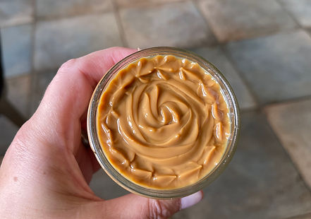 A hand holds an open jar filled with creamy Tres Leches Milk, swirled smoothly on top. The background features a tiled floor, suggesting a kitchen setting.