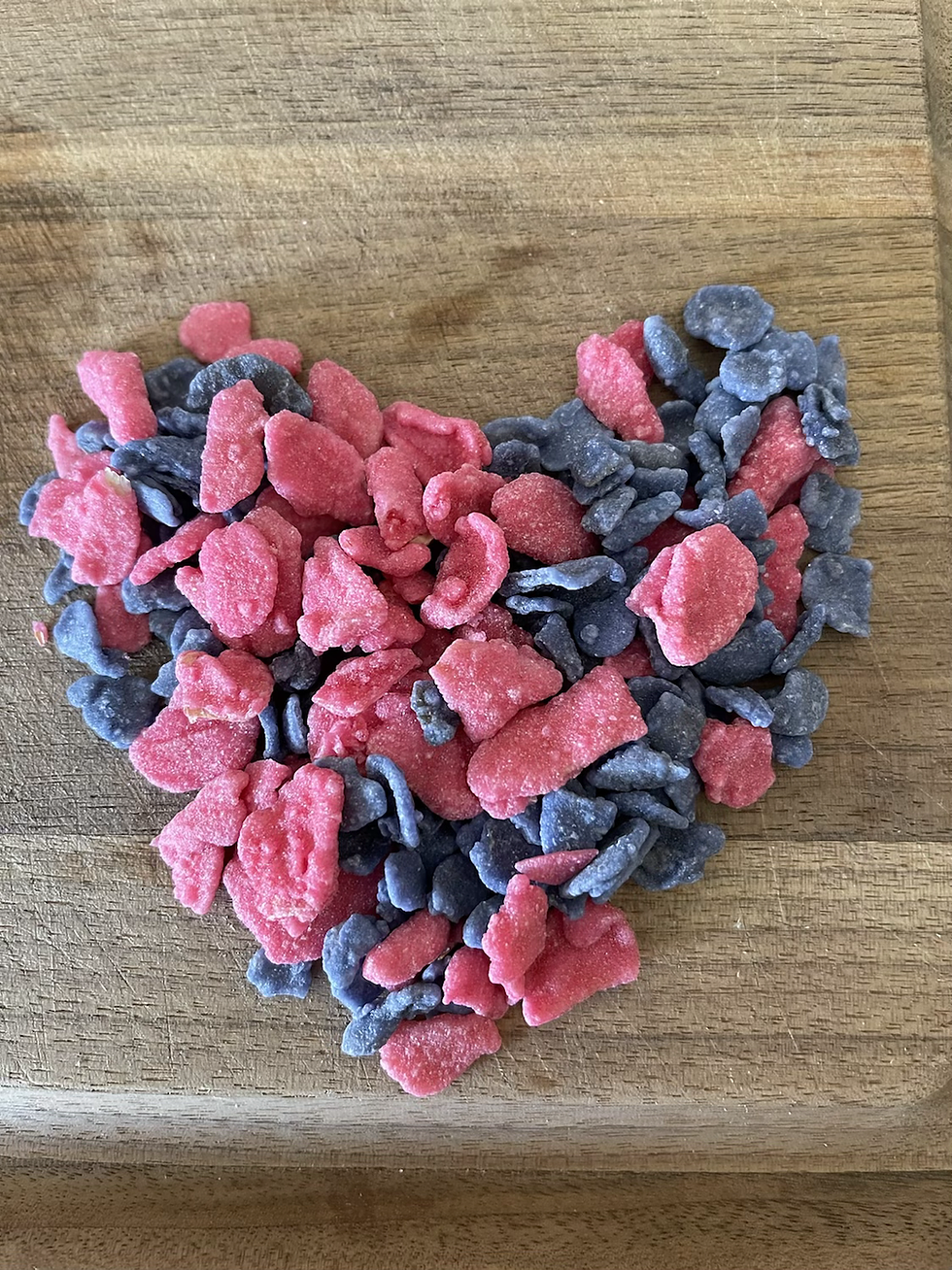 Red and blue colored candied flower petals shaped into a heart on a wooden surface. The candies have a frosted appearance, creating a playful mood.