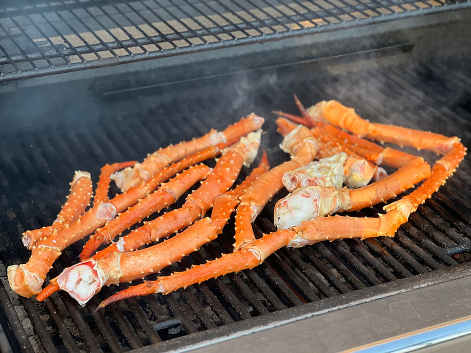 Red crab legs on a grill, emitting steam. The orange-pink color contrasts with the black grill, suggesting an outdoor cooking setting.