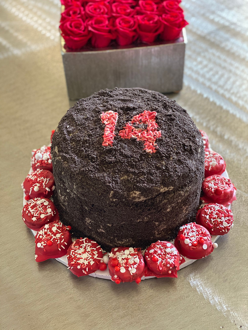 Chocolate cake with "14" in red icing, surrounded by red, sprinkle-topped cookies. Background shows red roses in a box.