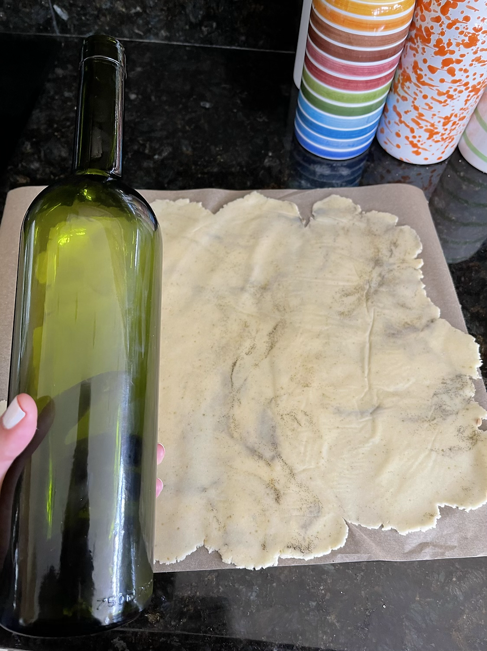 Hand holding a green wine bottle rolls out dough on a dark countertop. Colorful striped and speckled containers in the background.