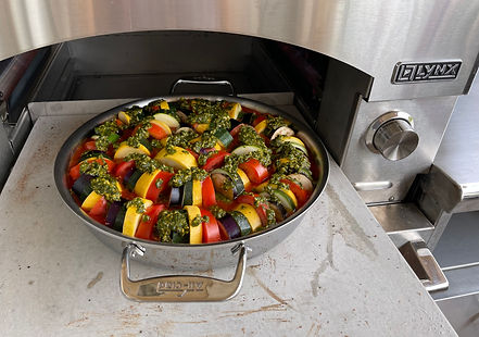 A vibrant tray of ratatouille with layered zucchini, squash, tomatoes, and eggplant, topped with green pesto, is cooking in a sleek silver oven.