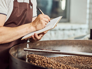 Coffee roaster in apron takes notes beside a drum full of freshly roasting beans in a workshop.