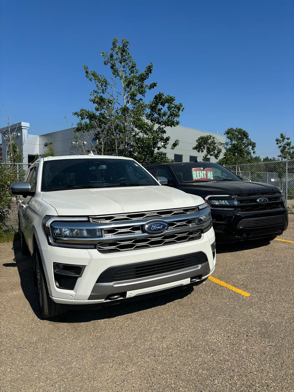 Two Ford SUVs 7 passenger vehicles, one white and one black, parked; "RENT ME" sign on black SUV. Clear blue sky, trees, and building in the background.