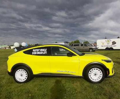 Yellow car with text "Fastest Vehicle Ever On Our Lot" on side, parked on grass under cloudy skies. White wheels, vehicles in background.
