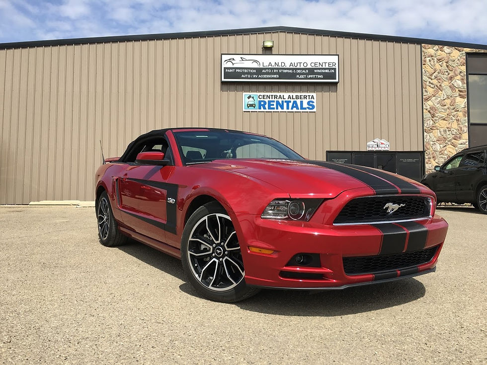 Red Mustang with black racing stripes parked outside L.A.N.D. Auto Center. Bright sunny day, Central Alberta Rentals sign on building.