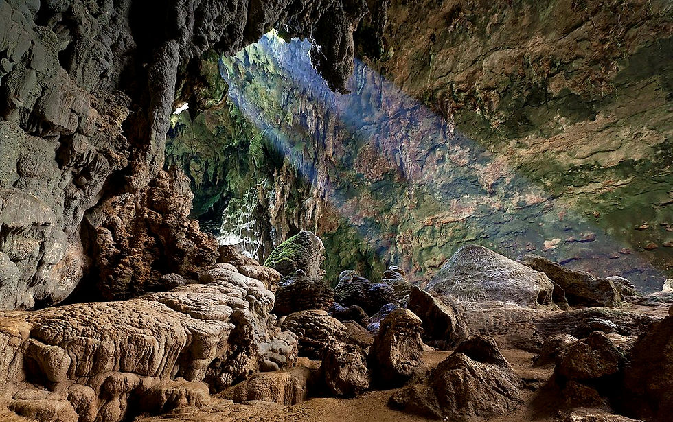 Looking at a ray of sunlight entering through opening the jagged rock celling of brown stone cave.