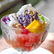 Colorful Halo- Halo dessert in a glass bowl featuring purple ice cream, coconut strips, mango, jelly, and sprinkled rice puffs on ice. Bright and vibrant.