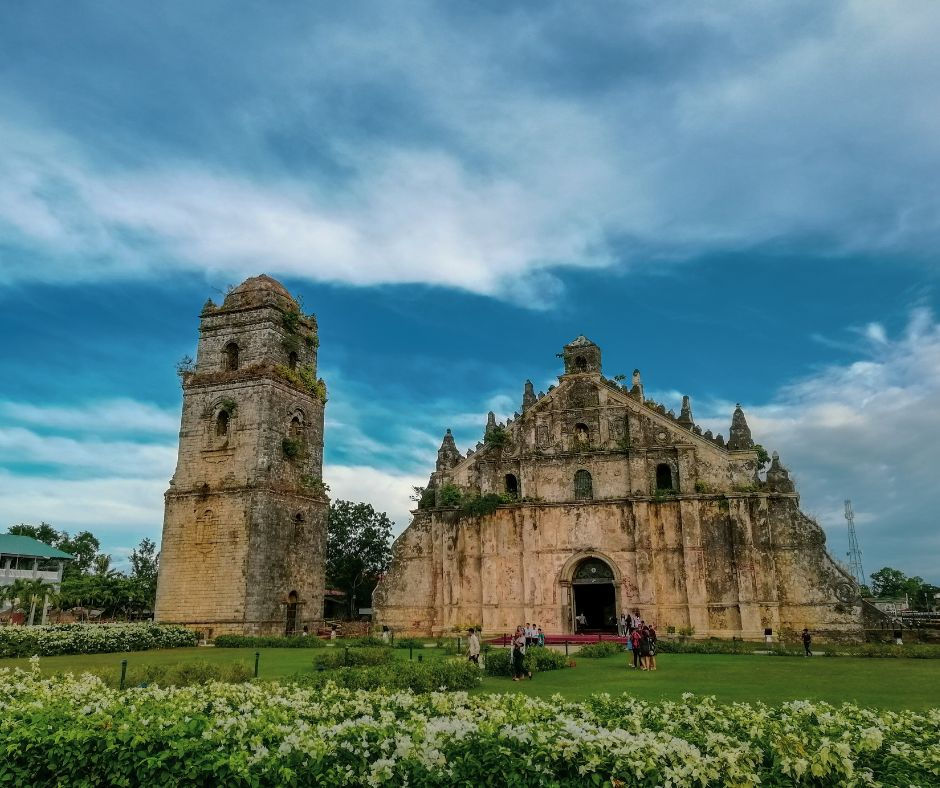 Historic stone church with a tall bell tower in a green garden. Groups of people explore the lush landscape under a clear blue sky.