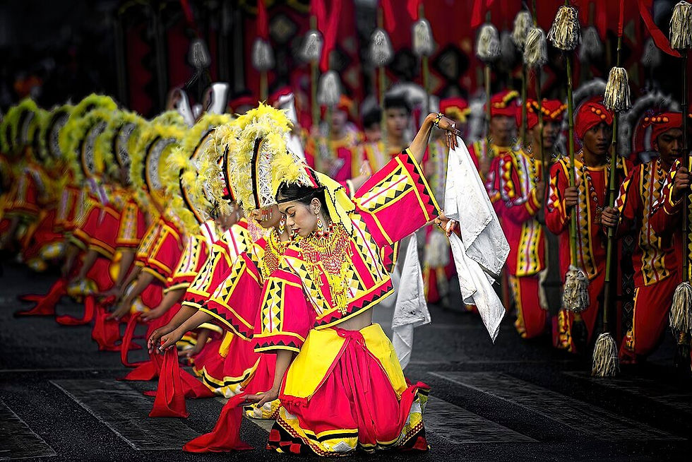 Kaamulan Festival dancers in vibrant yellow and red costumes perform in unison, displaying elaborate headdresses and expressive movements in a festive street setting.