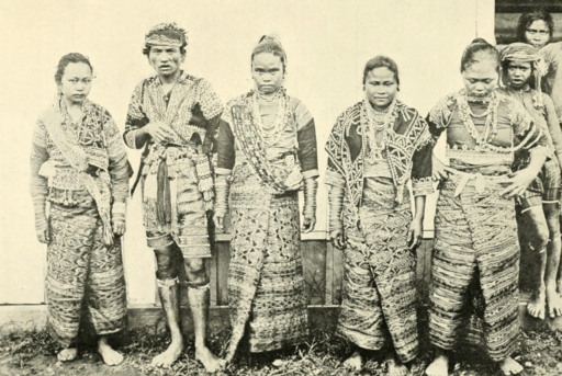 Bagobos in Davao, five people stand in traditional attire with intricate patterns. They pose against a wooden wall, exuding a solemn mood.