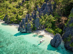 Aerial view of a tropical cove with turquoise water, limestone cliffs, palm trees, and a small boat by the white sand beach.
