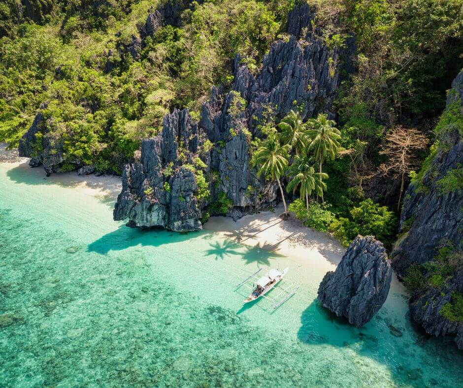 Aerial view of a tropical cove with turquoise water, limestone cliffs, palm trees, and a small boat by the white sand beach.