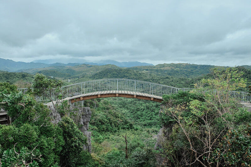 Masungi Georeserve Trail Bridge, a curved bridge spans a lush green valley under a cloudy sky, surrounded by dense trees and distant hills.