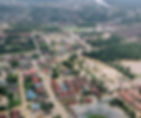 Aerial view of a flooded town, with submerged roads and buildings. Trees and rooftops are visible, surrounded by muddy water. Overcast mood.