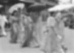 People in traditional attire walk on a sunny street, holding parasols. Background shows buildings and a vehicle with "Manila" visible.