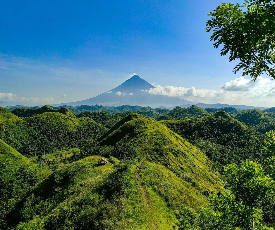 Lush green hills under a clear blue sky with a distant Mayon volcano in the background. A hut sits on a hilltop, surrounded by vibrant foliage.