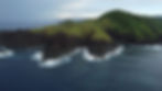 Rocky green island with lush vegetation surrounded by dark blue ocean waves under a cloudy sky. No text visible, serene atmosphere.