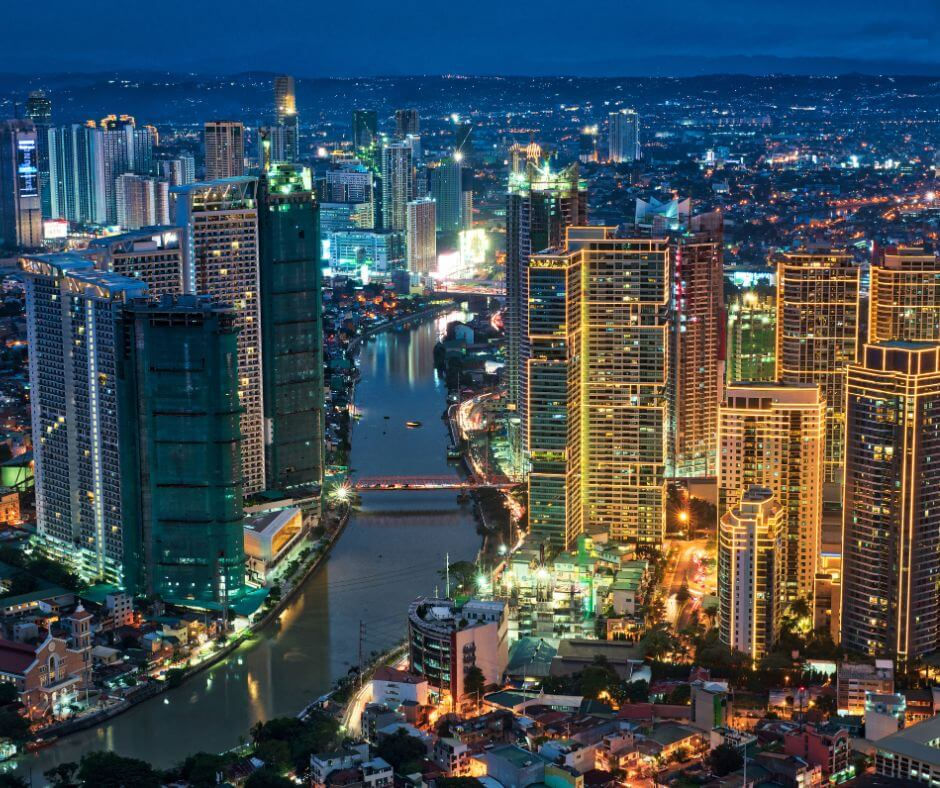 Manila City skyline at night with illuminated skyscrapers, a river running through, and bright lights reflecting on water, creating a vibrant scene.