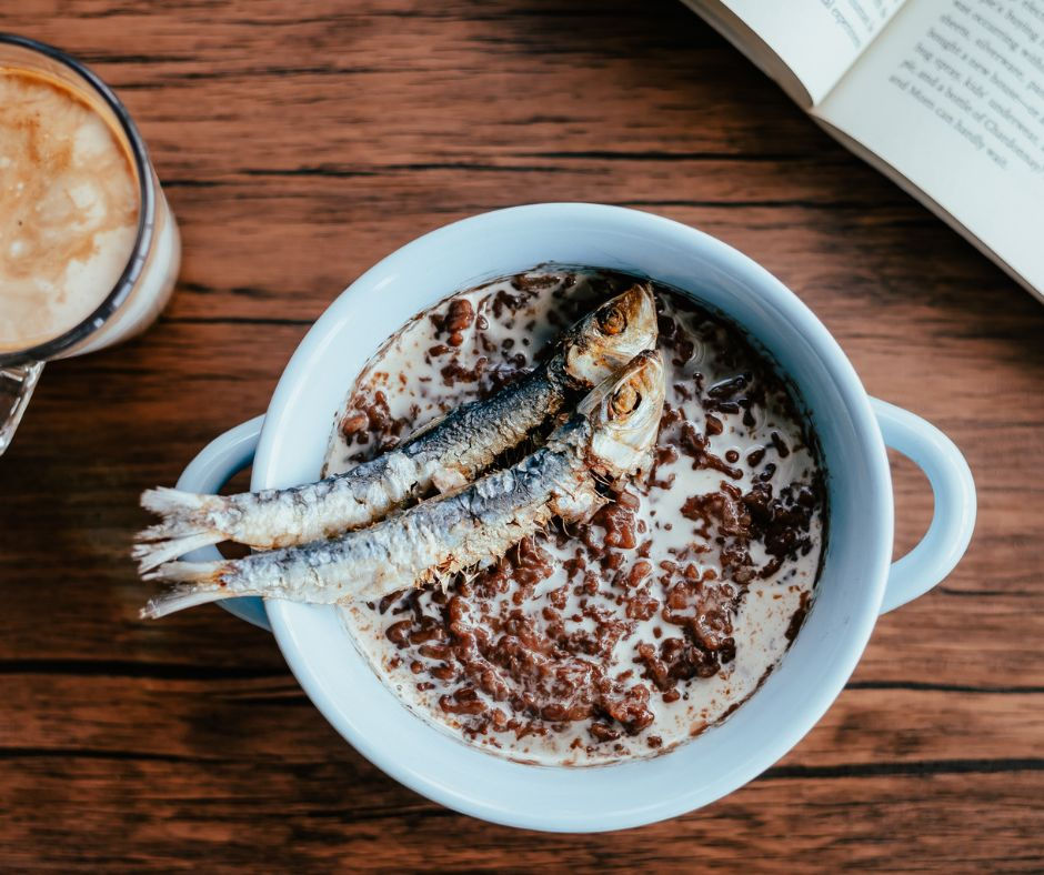 Blue bowl with chocolate cereal and two sardines on top, on wooden table. A coffee and open book are nearby, creating an unexpected mix.
