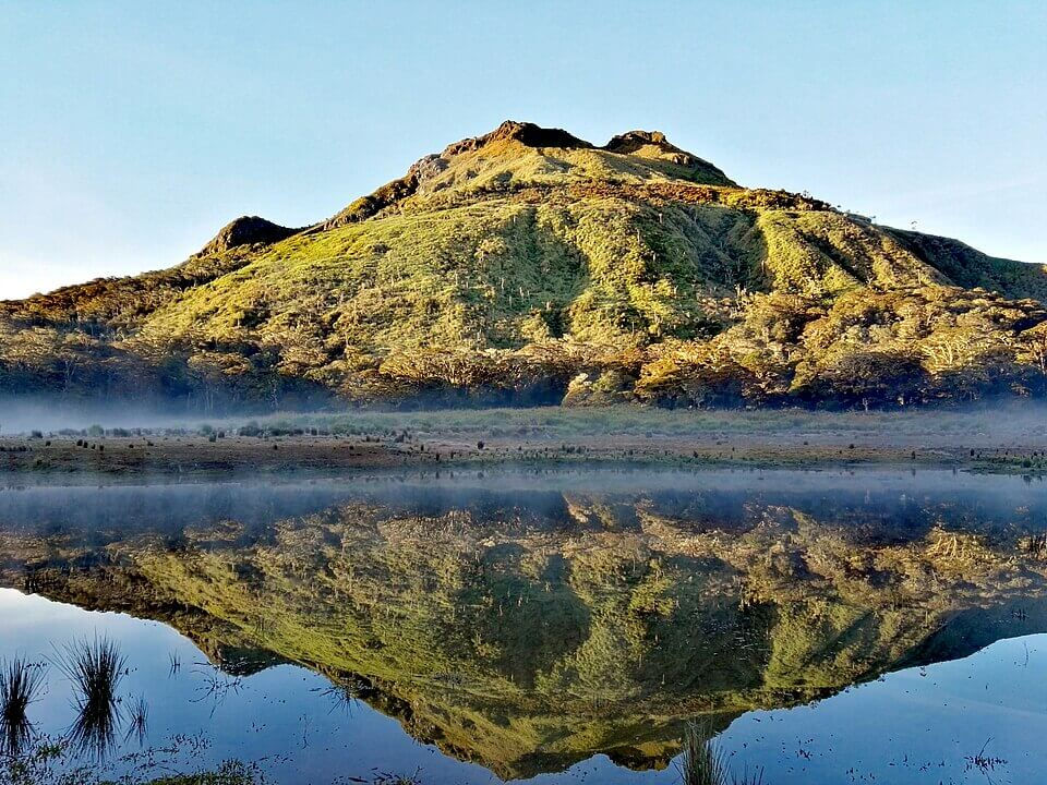 Green mountain reflected in a calm lake under a clear blue sky. The scene is peaceful, with light mist near the water's edge.