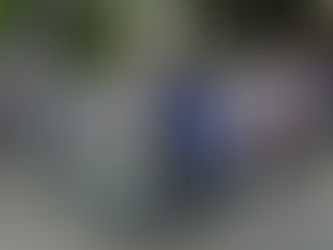 Men ride motorized tricycles in an urban street with trees and signs. The vehicles display colorful numbers and text, exuding a busy atmosphere.