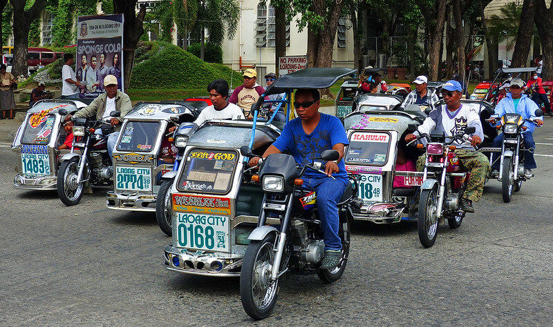 Men ride motorized tricycles in an urban street with trees and signs. The vehicles display colorful numbers and text, exuding a busy atmosphere.