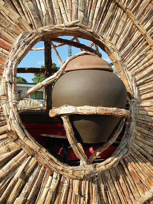 Woven straw frame surrounds a large Burnay clay pot in sunlight. Background shows partial trees and buildings, creating a natural, rustic scene.