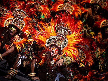 Masked dancers in orange-red feathered costumes shout and pose in a lively parade against a dark background.