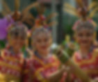 Three women in vibrant red traditional attire with beaded headdresses smile while holding decorated bamboo instruments, exuding joy.