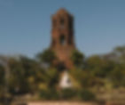 Old brick bell tower amidst lush greenery and clear sky, with a white statue at the base, creating a serene and historic atmosphere.