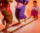 Two people dance between bamboo poles, wearing vibrant patterned clothing. One person kneels, holding the poles. Warm orange lighting.