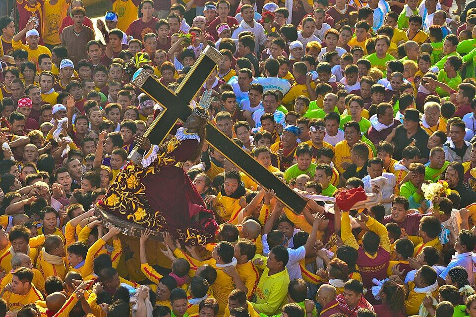 Crowd in yellow shirts carrying a religious statue with a cross during a procession. The scene is vibrant and energetic.