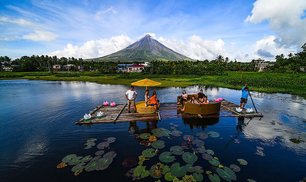 People relax on a floating raft with a yellow umbrella on a calm lake, lotus flowers nearby, and Mayon volcano in the background under a blue sky.