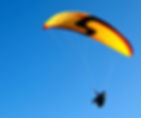 Person paragliding with a yellow and red wing against clear blue sky. The paraglider’s shape forms an arc, creating a sense of freedom.