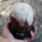 Asin Tibuok, a hand holds a cracked, egg-shaped object with a white top and brown base. Pebbled ground in the blurred background. Natural texture visible.