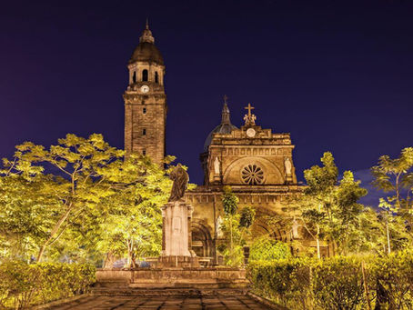 Illuminated Manila Cathedral and statue at night, ornate architecture with a large clock tower. Trees and a clear dark blue sky enhance the scene.