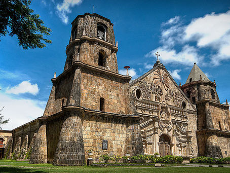 Historic stone Miagao Church in Iloilo with ornate facade, set against a blue sky with clouds. Green lawn and trees frame the rustic structure. Calm atmosphere.