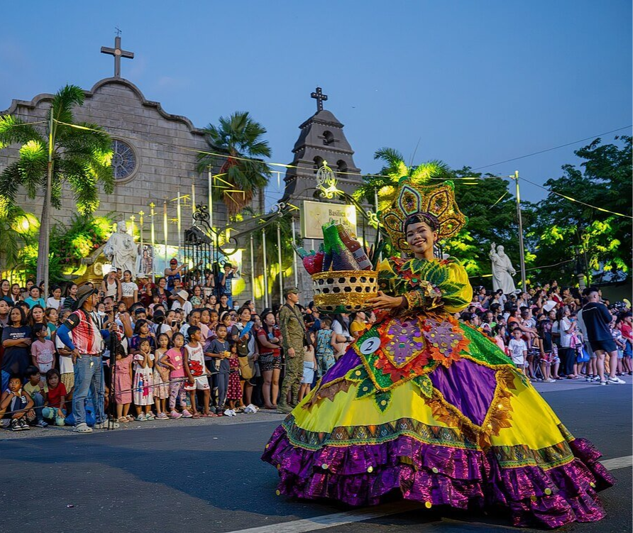 Colorful festival parade with a smiling person in vibrant attire holding a basket. Crowd watches by a church under a clear evening sky.