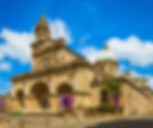 Historic stone cathedral with statues and a clock tower under a bright blue sky. Purple banners adorn the facade. Peaceful and grand setting.