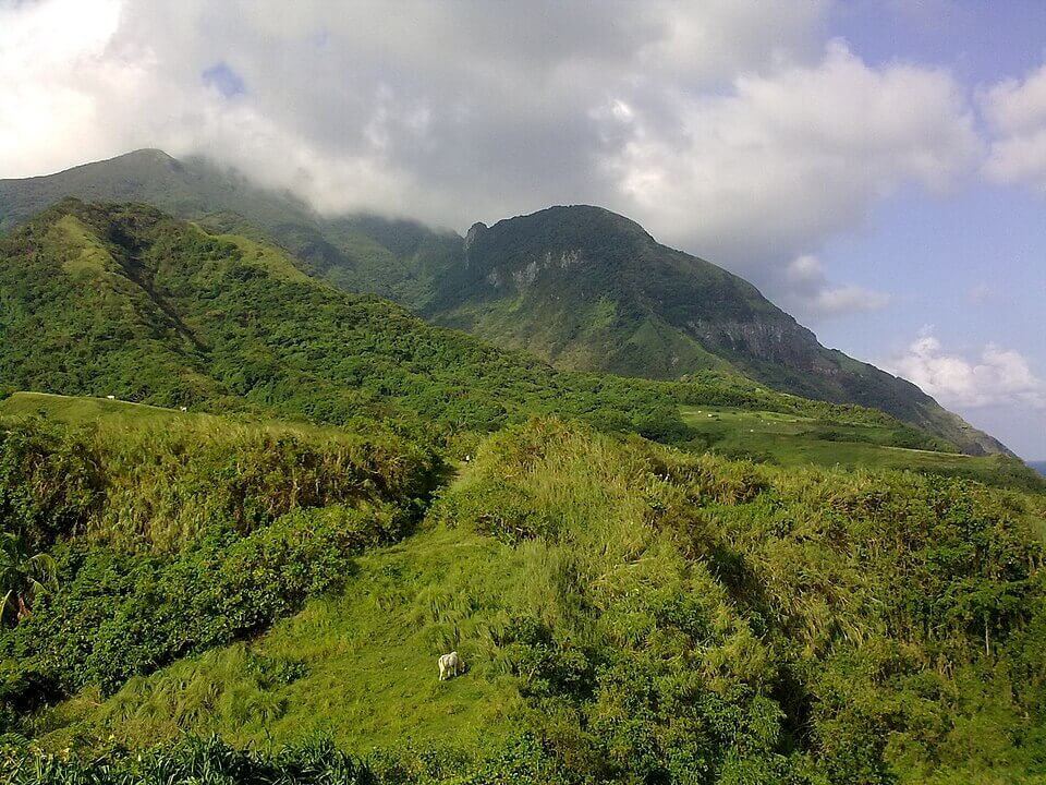 Lush green mountains under cloudy sky, with a small goat grazing on a grassy hillside.